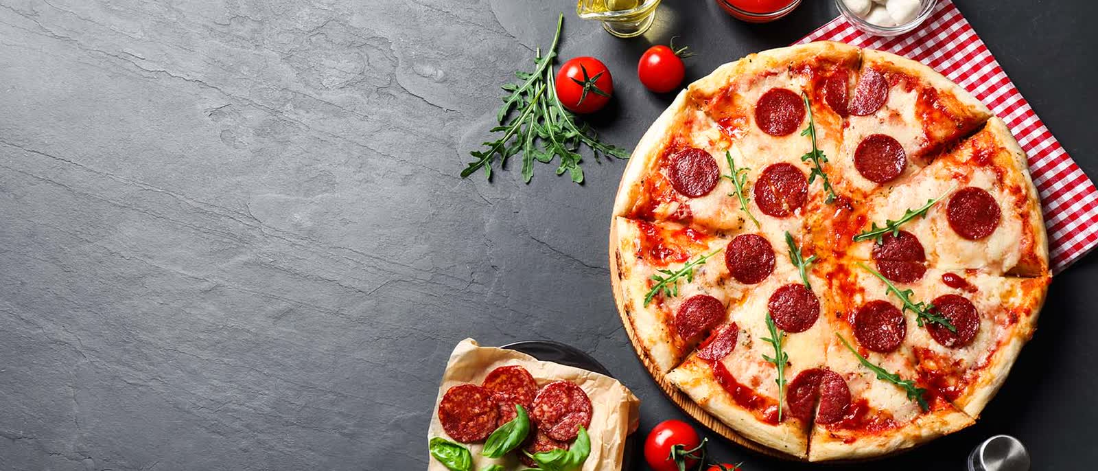 Een pizza met pepperoni en rucola op een houten plank, op leisteen, met tomaten, mozzarella en tomatensaus ernaast.
